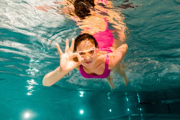 Cheerful woman in googles showing ok sign while diving underwater — Stock Photo