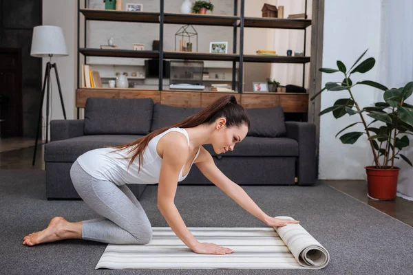 Hermosa joven rodando alfombra de fitness para practicar yoga en casa - foto de stock