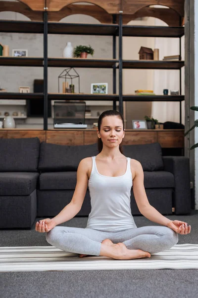 Beautiful young woman sitting in lotus pose and meditating at home in living room — Stock Photo