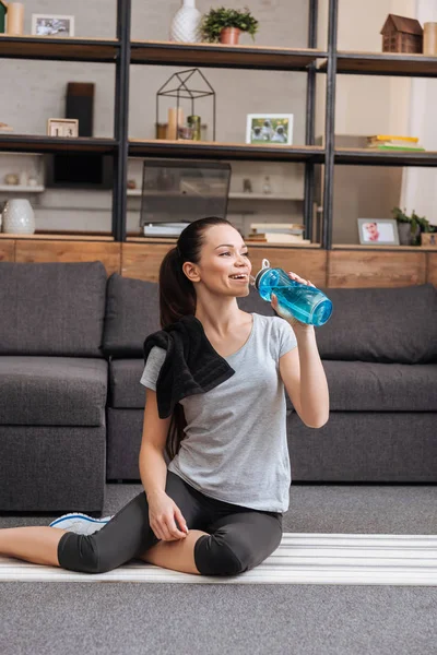 Sportlerin sitzt auf Fitnessmatte und trinkt Wasser aus Sportflasche im Wohnzimmer — Stockfoto