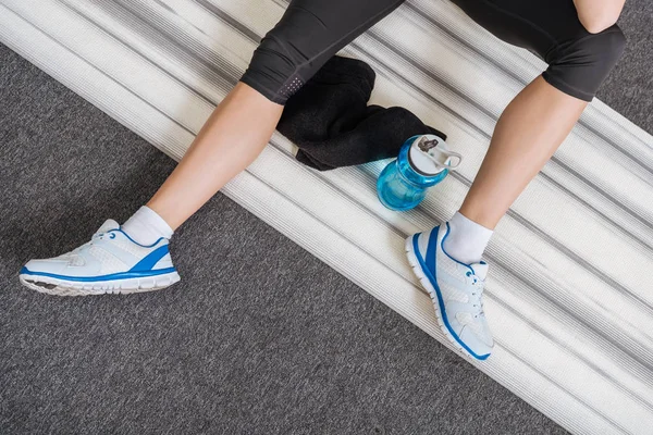 Vue recadrée d'une sportive assise sur un tapis de fitness avec bouteille de sport et serviette à la maison — Photo de stock