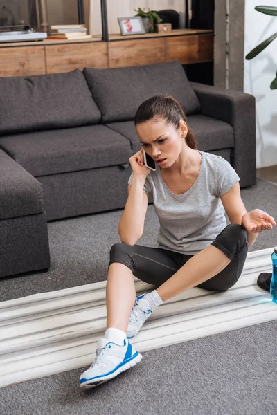 Frustrated sportswoman sitting on fitness mat and talking on smartphone at home — Stock Photo