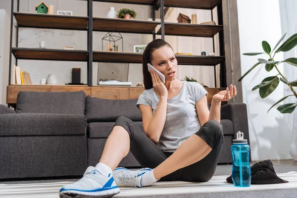 Deportista frustrado sentado en la alfombra de fitness y hablando en el teléfono inteligente en casa - foto de stock