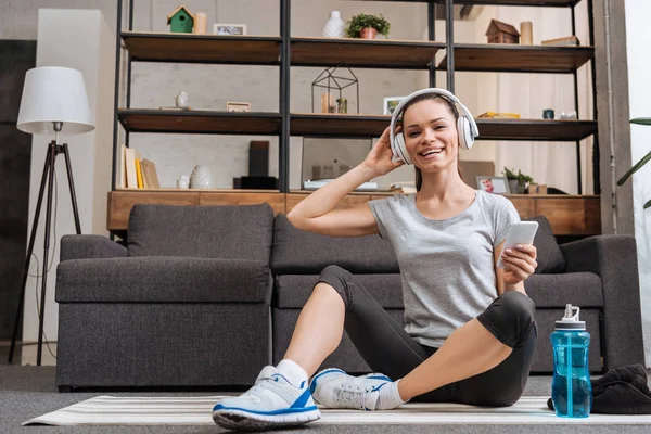 Lächelnde Sportlerin mit Kopfhörern, die sitzt, in die Kamera schaut und zu Hause das Smartphone benutzt — Stockfoto