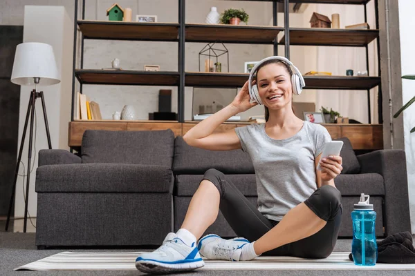 Sorrindo esportista em fones de ouvido sentado, olhando para a câmera e usando smartphone em casa — Fotografia de Stock