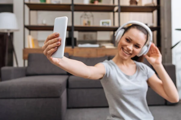 Belle femme souriante dans les écouteurs prendre selfie sur smartphone à la maison — Photo de stock