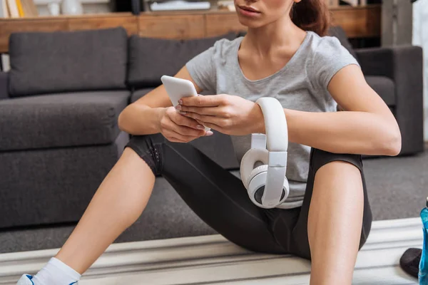 Vista ritagliata di sportiva con cuffie utilizzando smartphone a casa — Foto stock