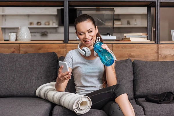 Привлекательная улыбающаяся спортсменка с наушниками, сидящая на диване и использующая смартфон дома — стоковое фото