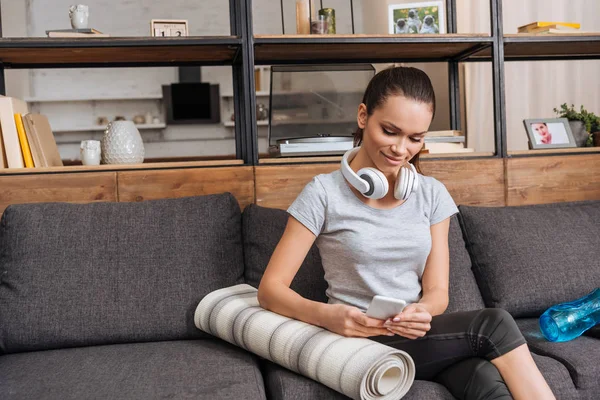 Attractive sportswoman with headphones sitting on couch and using smartphone at home — Stock Photo