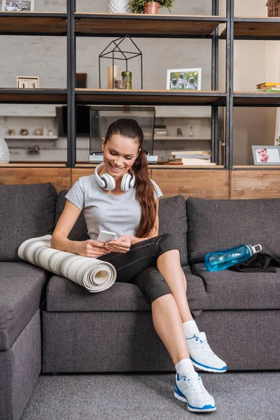 Attraente sportiva sorridente con cuffie seduta sul divano e con smartphone a casa — Foto stock