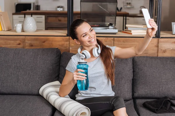 Belle sportive souriante avec écouteurs prenant selfie sur smartphone à la maison — Photo de stock