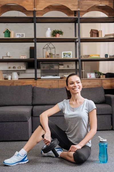 Lächelnde Sportlerin mit Bauchtrainer und Sportflasche zu Hause — Stockfoto