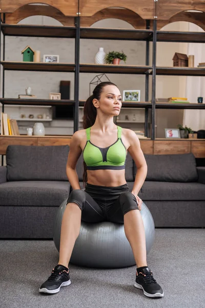 Beautiful sportswoman sitting on fitness ball at home and looking away — Stock Photo