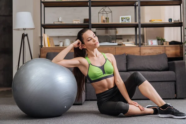 Tired sportswoman sitting near fitness ball at home and looking away — Stock Photo