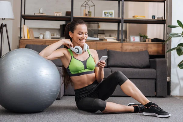 Sportiva sorridente in cuffia seduta con palla fitness e con smartphone a casa — Foto stock