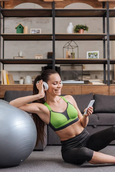 Smiling sportswoman in headphones sitting with fitness ball and using smartphone at home — Stock Photo