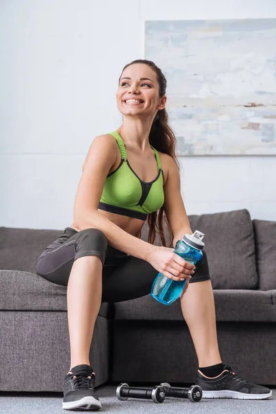 Smiling sportswoman sitting with sport bottle on couch at home — Stock Photo