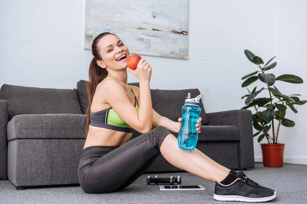 Smiling sportswoman holding water bottle and eating apple at home — Stock Photo