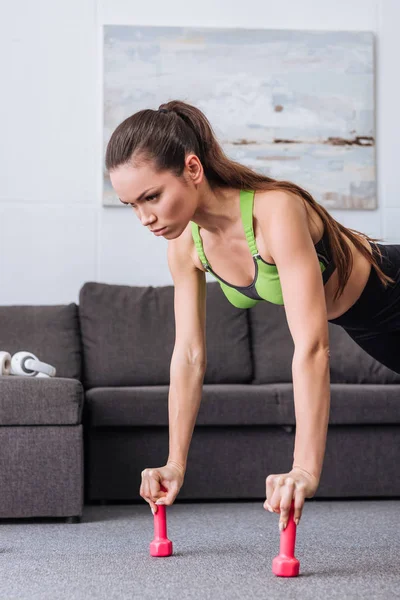 Focused sportswoman doing push ups with dumbbells at home — Stock Photo