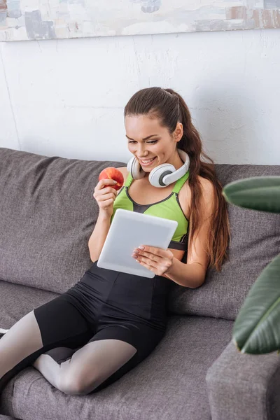 Deportista sonriente con auriculares sosteniendo manzana y usando tableta digital en casa - foto de stock