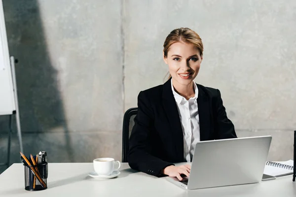 Smiling young businesswoman using laptop and looking at camera in office — Stock Photo