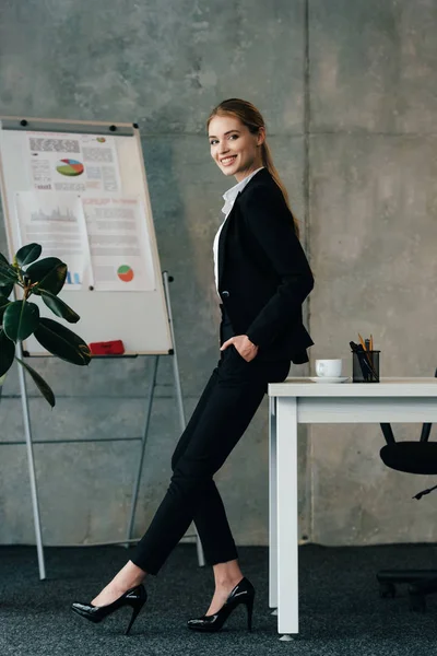 Smiling businesswoman standing by work desk and keeping hands in pockets — Stock Photo