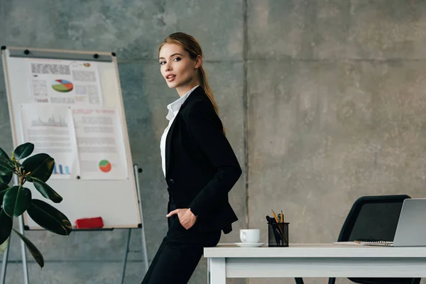 Beautiful businesswoman standing by desk with hands in pockets — Stock Photo