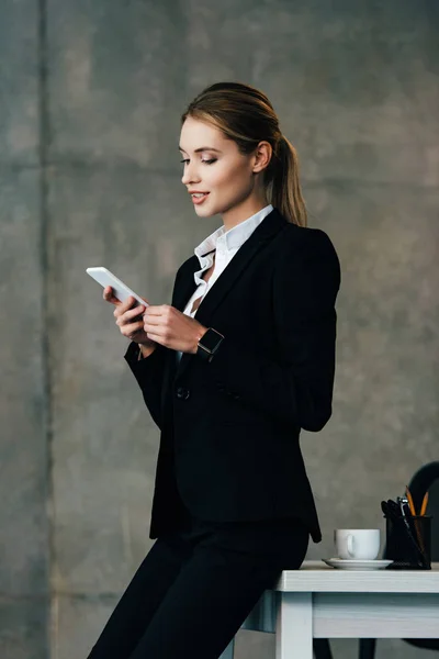 Smiling businesswoman standing by desk and using smartphone in office — Stock Photo