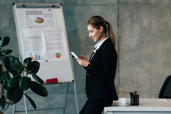 Smiling businesswoman standing by work table and using smartphone — Stock Photo