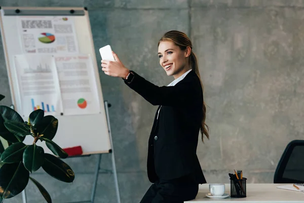 Femme d'affaires souriante prenant selfie tout en se tenant près du lieu de travail dans le bureau — Photo de stock