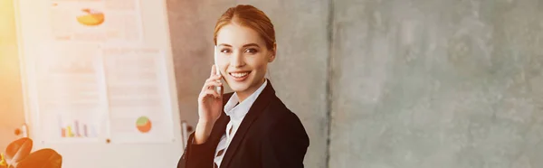 Sonriente mujer de negocios hablando en el teléfono inteligente y mirando a la cámara en la oficina - foto de stock