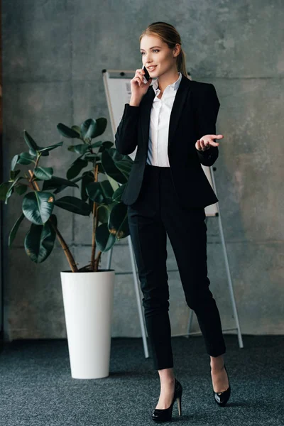 Привлекательная улыбающаяся деловая женщина разговаривает по телефону, стоя возле большого цветочного горшка — стоковое фото