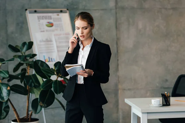 Junge seriöse Geschäftsfrau spricht mit Smartphone und hält Notizbücher in der Hand — Stockfoto