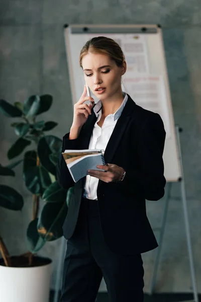 Young businesswoman seriously talking on smartphone while holding notebooks in hands — Stock Photo