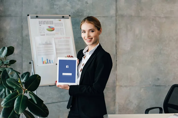 Pretty smiling businesswoman holding digital tablet with facebook on screen — Stock Photo