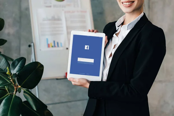 Vista parcial de mujer de negocios bastante sonriente sosteniendo tableta digital con facebook en la pantalla - foto de stock