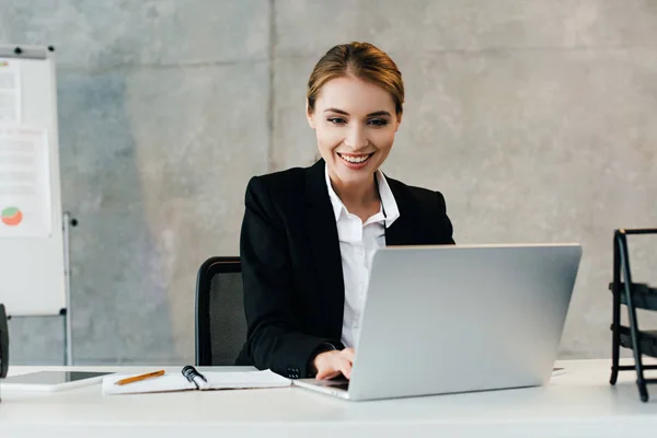 Mujer de negocios sonriente sentada en el lugar de trabajo y usando computadora portátil - foto de stock
