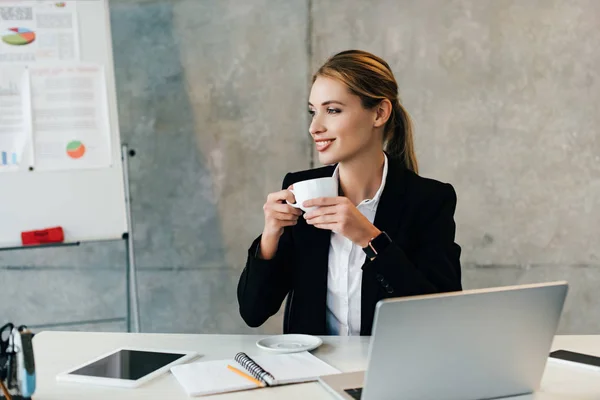Гарненька усміхнена бізнес-леді сидить на робочому місці і п'є каву — Stock Photo