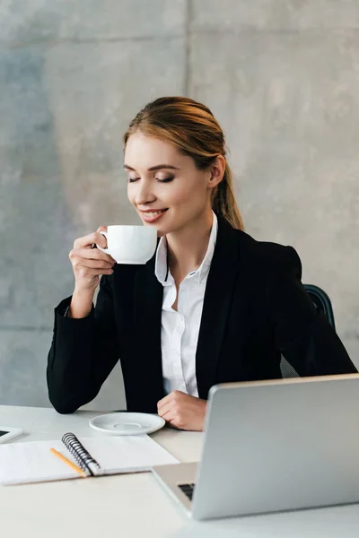 Smiling businesswoman enjoys drinking coffee with closed eyes while sitting at workplace in office — Stock Photo