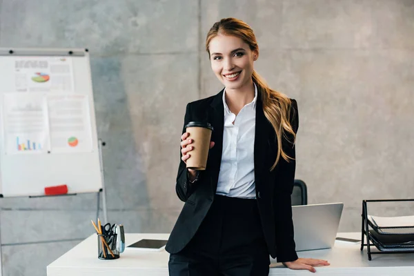 Beautiful smiling businesswoman standing by workplace with disposal cup of coffee — Stock Photo