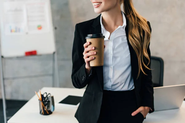 Обрезанный вид на улыбающуюся деловую женщину, остановившуюся за рабочим столом с чашкой кофе — стоковое фото