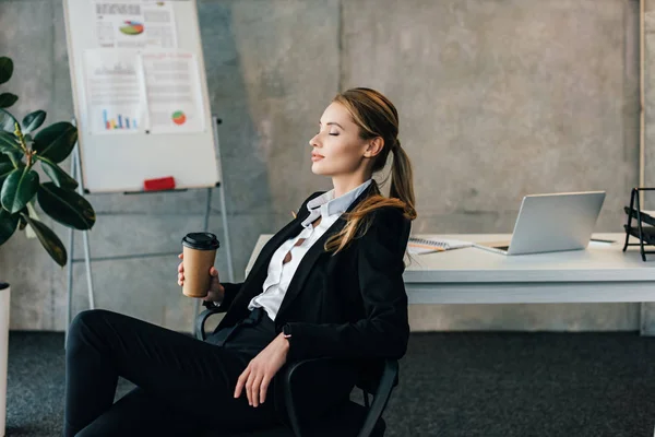 Jeune femme d'affaires assise sur une chaise aux yeux fermés et tenant une tasse de café en papier — Photo de stock
