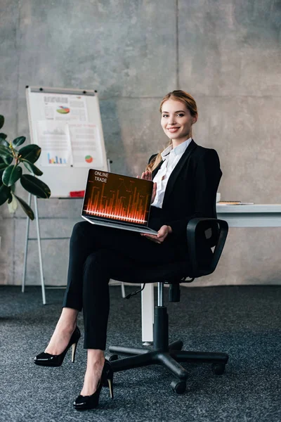 Sonriente joven mujer de negocios sentado en la silla y la celebración de la computadora portátil con las tasas de comercio en línea en la pantalla — Stock Photo