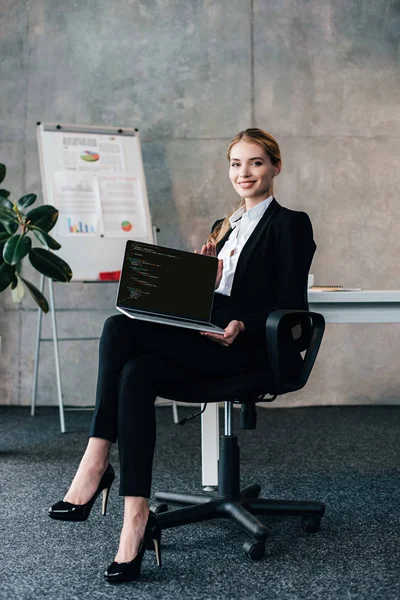Lächelnde Geschäftsfrau sitzt auf Stuhl und hält Laptop mit Programmiercode auf dem Bildschirm — Stockfoto