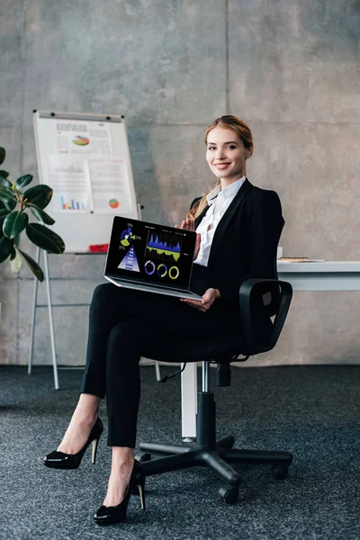 Mujer de negocios sonriente sentada en la silla y sosteniendo el ordenador portátil con diagramas comerciales en la pantalla - foto de stock