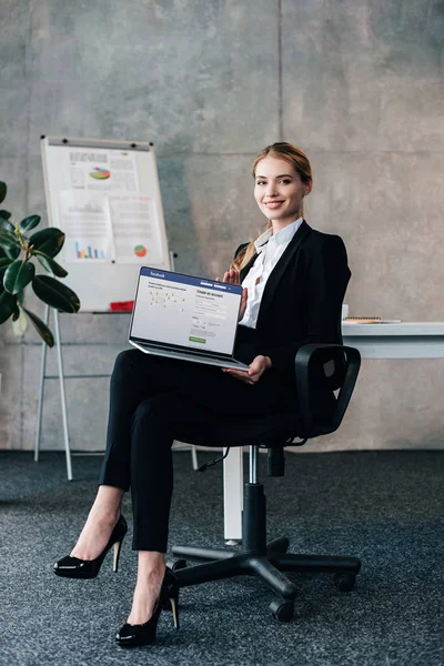 Lächelnde Geschäftsfrau sitzt im Stuhl und hält Laptop mit Facebook auf dem Bildschirm — Stockfoto