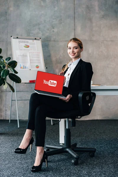 Sonriente mujer de negocios mirando a la cámara y la celebración de ordenador portátil con youtube sitio web en el monitor - foto de stock