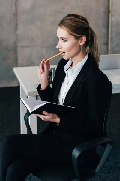 Selective focus of beautiful businesswoman holding notebook and gnawing pencil — Stock Photo