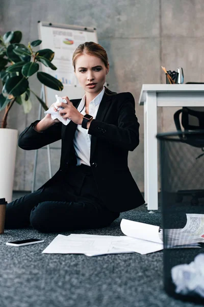 Businesswoman sitting on floor and throwing crumpled papers to basket — Stock Photo