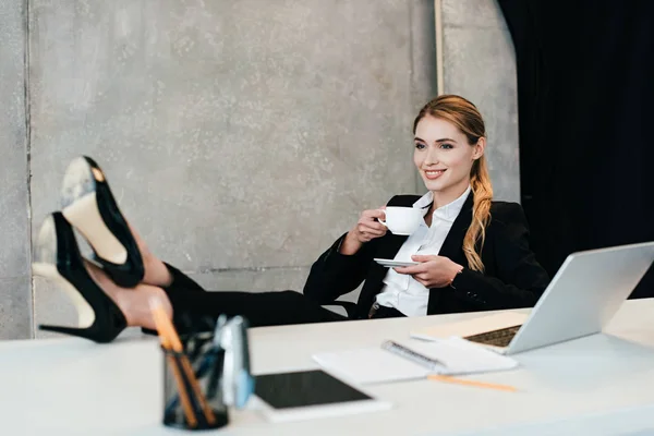 Selective focus of beautiful woman with legs on table sniling and drinking coffee — Stock Photo